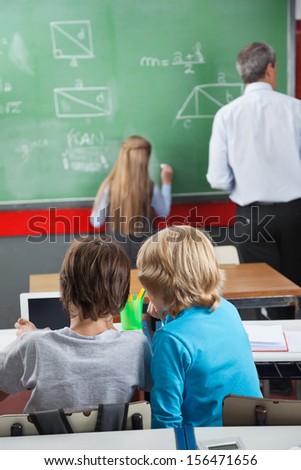 Rear view of little schoolboys using digital tablet at desk with classmate and teacher in foreground