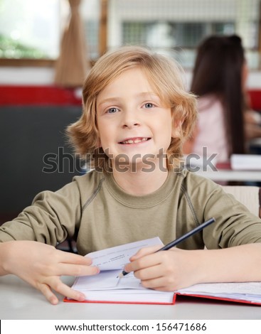 Portrait of little schoolboy writing in book at desk with classmate in background