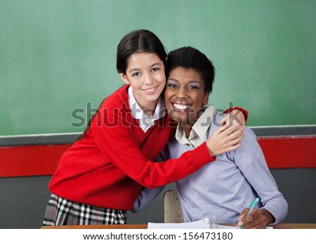 Portrait of happy schoolgirl hugging professor at desk in classroom
