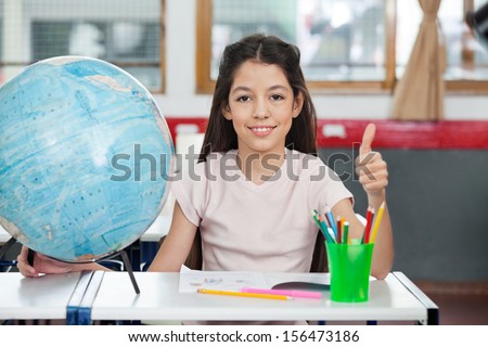 Portrait of cute schoolgirl gesturing thumbs up while sitting with globe and organizer at desk in classroom