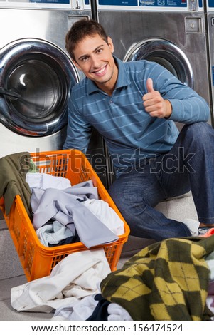 Portrait of young man gesturing thumbs up with dirty clothes on floor at laundry