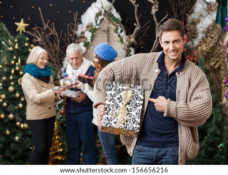 Portrait of happy young man pointing at Christmas present with family standing in background at store