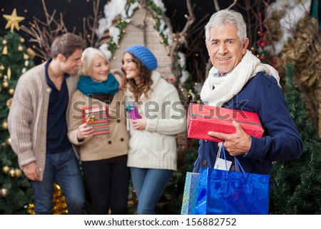 Portrait of happy senior man holding Christmas presents with family standing in background at store