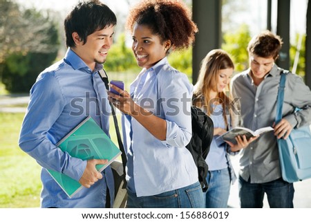 Happy multiethnic friends with mobilephone and book looking at each other in college campus
