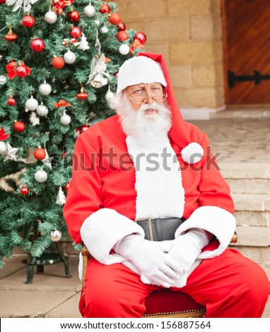 Senior man dressed as Santa Claus sitting in front of Christmas tree outside house