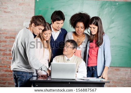 Mature male teacher with laptop explaining lesson to group of students at desk in classroom