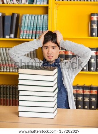 Confused young man with head in hands looking at stacked books in college library