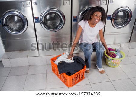 Portrait of young African American woman with baskets of dirty clothes sitting against washing machines