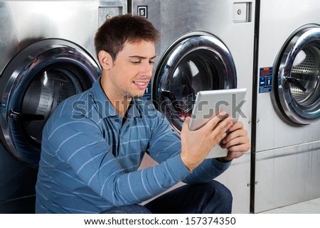 Young man using digital tablet while sitting against washing machines at laundry