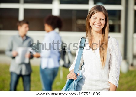 Portrait of happy young woman carrying shoulder bag with students in background on university campus