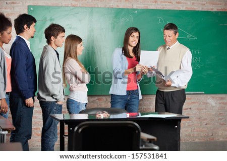Happy female student and teacher looking at test result with classmates standing in classroom