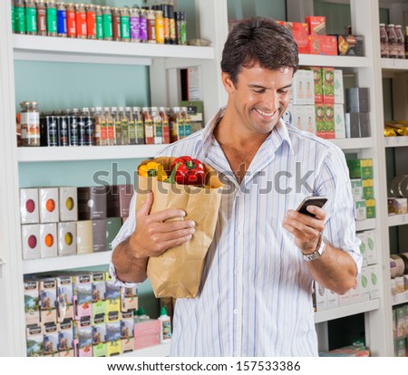 Happy Male Customer With Grocery Paper Bag Using Mobile Phone In Supermarket