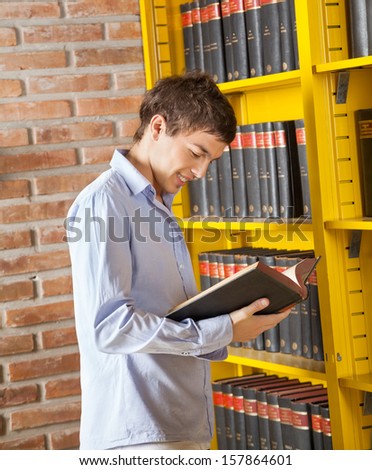 Young male student reading book while standing by shelf in college library