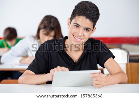 Portrait of teenage schoolboy using digital tablet at desk with classmates studying in background