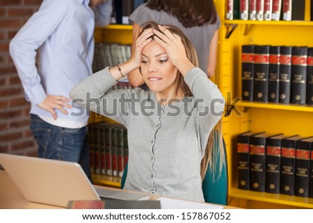 Worried young woman biting lip while looking at laptop in college library
