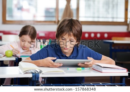 Elementary schoolboy using digital tablet with classmate studying in background at classroom