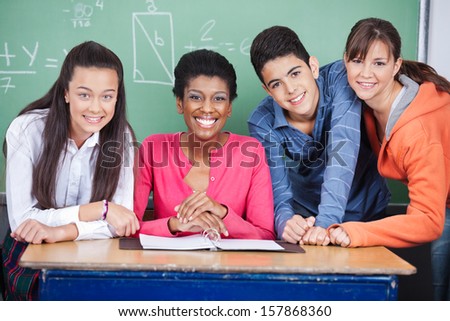 Portrait of happy young teacher with teenage students at desk in classroom