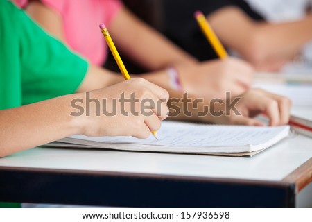 Midsection Of Teenage Classmates Writing In Book At Desk