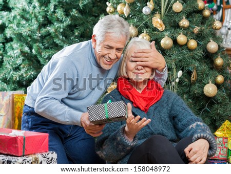 Senior man covering woman\'s eyes while surprising her with Christmas gifts in store