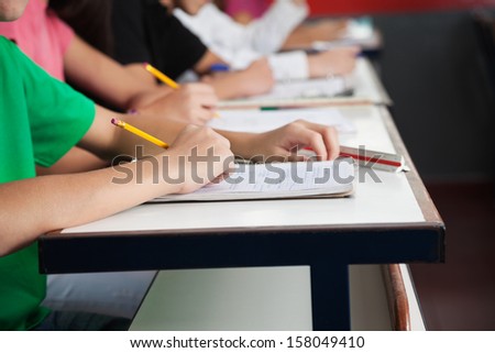 Midsection of high school students writing on paper at desk in classroom