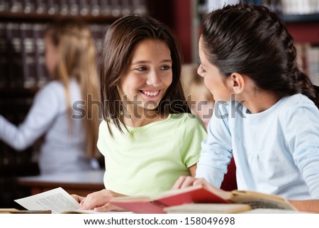 Happy little schoolgirl looking at female friend while studying in library