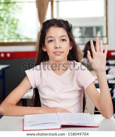 Little schoolgirl raising hand while sitting at desk in classroom