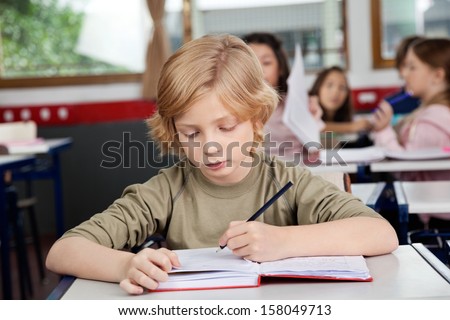 Cute schoolboy writing in book at desk with classmates in background