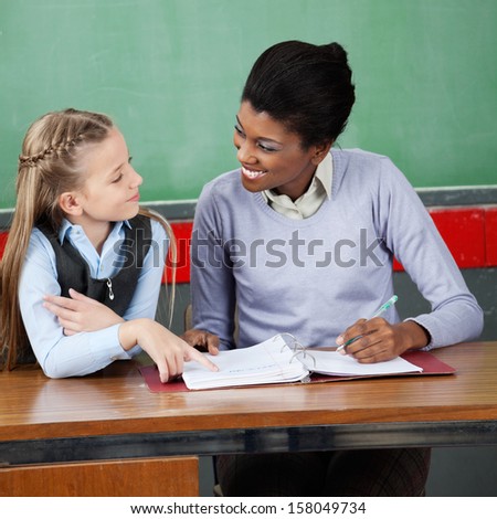 Happy young female professor looking at little schoolgirl at desk in classroom