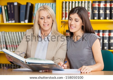 Happy student and teacher reading book together at table in university library