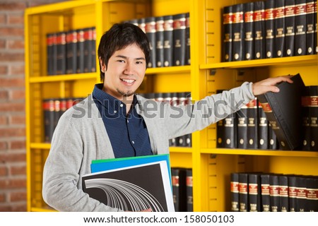 Portrait of happy male student choosing book from shelf in college library
