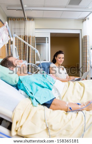 Portrait of young woman with laptop sitting by patient in hospital room