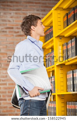 Side view of young male student holding books while looking at shelf in college library