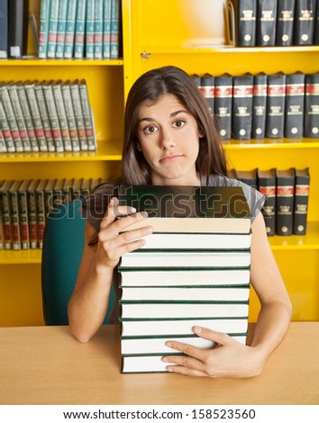 Portrait of confused female student with stacked books sitting at table in college library