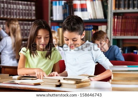 Little Schoolgirls Reading Book Together While Sitting At Table In Library With Classmates In Background