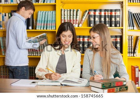 Happy young female friends studying together at table in university library