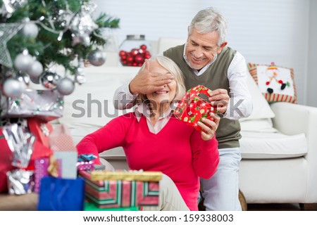 Happy senior man covering woman\'s eyes while giving Christmas gift at home