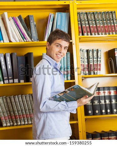 Side view portrait of happy male student with book standing against shelf in university library