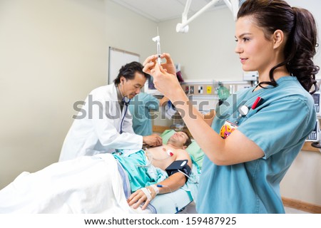Beautiful nurse preparing injection while doctor and colleague examining critical patient in hospital