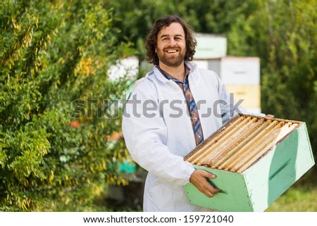 Portrait of happy male beekeeper carrying crate full of honeycombs at apiary