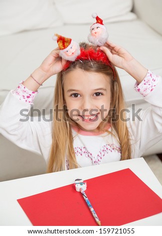 Portrait of happy girl with pencil and cardpaper holding Santa headband at home