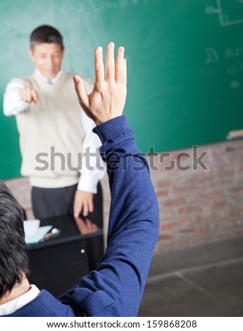 Rear view of male student raising hand to answer question in classroom