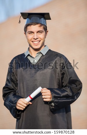 Portrait of happy young man in graduation gown holding certificate on university campus