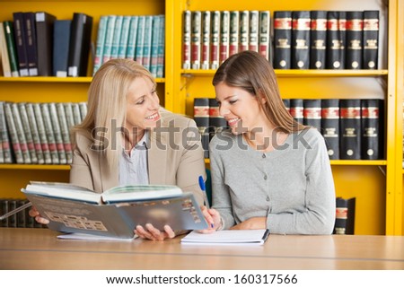 Happy university teacher holding book while explaining student in library