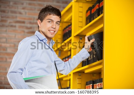Portrait of confident male student taking book from shelf in library