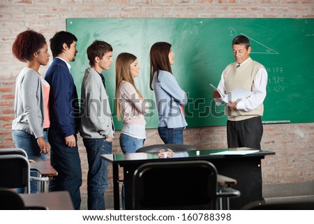 Mature male professor with exam results while students standing in a row at classroom desk