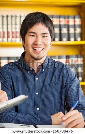 Portrait of happy male student sitting against bookshelf in university library