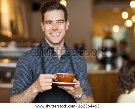 Portrait Of Happy Waiter Holding Coffee Cup While Standing In Cafeteria