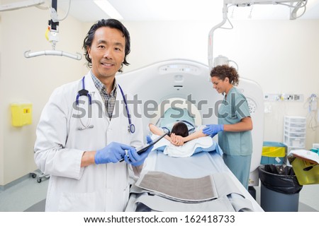 Portrait of mature doctor holding digital tablet while nurse preparing patient for CT scan in hospital