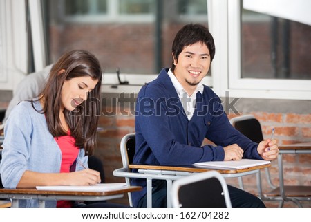 Portrait of happy college student sitting at desk with classmate in classroom