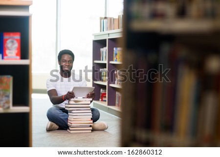 Full length portrait of young male student with digital tablet and books sitting on floor at library
